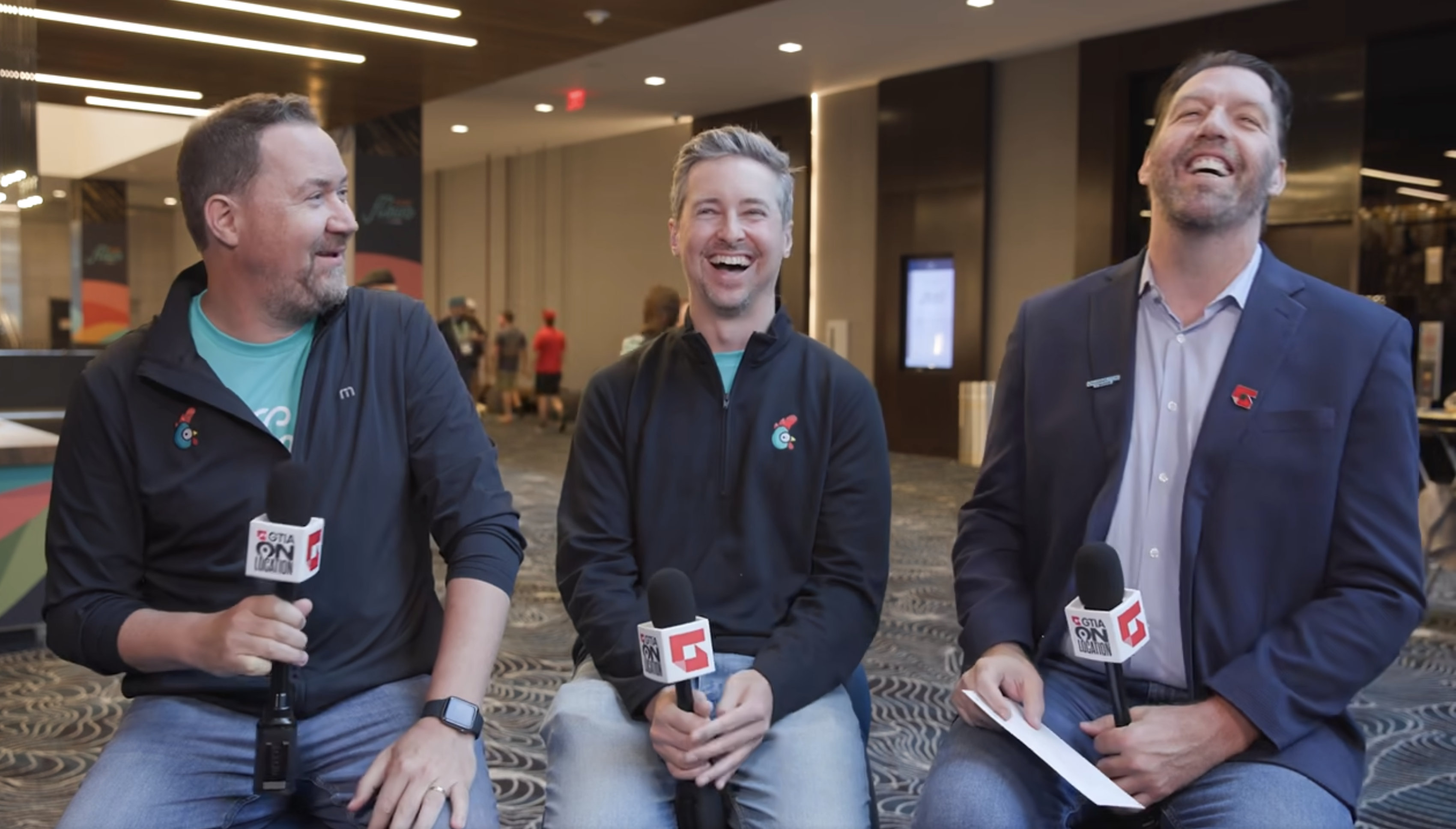Three attendees laughing mid-interview at an industry event, mics in hand.
